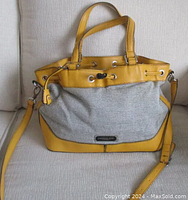 Front view of London Fog tweed and mustard yellow leather handbag with top handles and crossbody strap on a beige sofa.