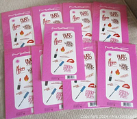 Nine sealed MAC promotional card and sticker sets laid out showing front side of pink packaging with makeup images and themed words.