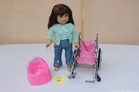 Full view of American Girl doll articulated standing next to wheelchair and pink potty chair on a white surface with beige background