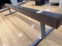 Side angled view of the gymnastics balance beam showing the brown suede-like covering and metal base stands.
