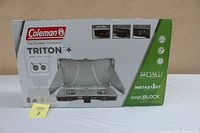 Front view of the Coleman Triton+ camping stove box showing the product image and main features such as high BTU, wind block, and instant start.