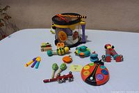Various children's musical instruments arranged on a white surface, including a mini drum with strap, wooden bee rattle, ladybug xylophone, maracas, tambourines, whistle, and toy flutes.