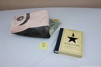 Photo showing the folded Hamilton-themed blanket next to the Hamilton The Revolution hardcover book closed on a white surface.