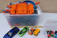 Plastic bin filled with orange and blue Hot Wheels race track pieces and assorted toy cars in storage boxes beneath