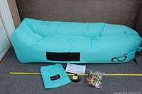 Turquoise Live Infinifty inflatable lounger fully inflated, showing size with measuring tape, next to turquoise storage bag, instruction booklet and bag of pool toys.