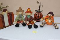 Full view of all five items: one wooden pumpkin, three scarecrow dolls in various fall-themed outfits, and one metal jack-o-lantern with witch hat.