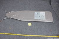 Full view of the new ironing board wrapped in plastic, lying on a carpeted floor with measuring tape indicating length.