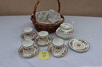 Full view of the tea set including tea pot, cups, saucers, small plates, and basket. White porcelain with multi-color floral and gold trim.