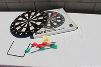 Photo showing magnetic dartboard, 8 darts in red and green, and metal stand on a white surface with packaging box in background labeled 'Safe Dart Set'