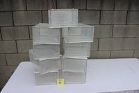 Stack of ten plastic shoe organizing boxes arranged in two pyramidal columns on a white surface in front of a grey brick wall.