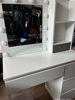 Photo of white vanity table showing lighted mirror with bulbs on frame, one drawer and open shelf with black fabric bin.