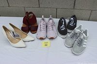 Photo shows six pairs of women's shoes arranged in two rows on a white surface against a gray cinder block wall. Included are white heels, burgundy sandals, pink running shoes, and multiple casual sneakers from Adidas and Vans.