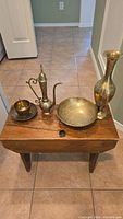 Five brass decor items on a wooden bench: vase, teacup with saucer, dish, Aladdin lamp, candle snuffer.