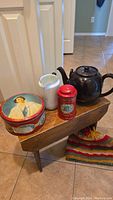Group photo of all four items: vintage Queen cookie tin, white china pitcher, red tin, and Brown Betty teapot with cozy on small wooden bench.