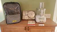 KitchenAid food processor with black patterned appliance cover, plastic tray holding attachments, and instruction booklet.