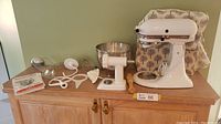 Overview of stand mixer, bowls, attachments, cover and booklet arranged on cabinet