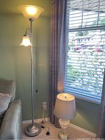Tall standing floor lamp with a frosted glass bowl shade on top and a smaller reading light attached, next to a sofa and window with blinds and curtains.