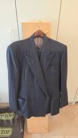 Front view of the dark wool double-breasted suit jacket with peak lapels hanging on a wooden hanger.