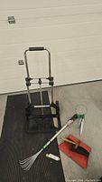 Photo showing collapsible hand trolley, small garden rake, dustpan, and brush on a mat in front of garage door.