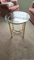 Round glass topped side table with brass finished legs and steel frame, photographed in a room with two armchairs and carpet.