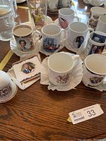 Top view of mugs and glassware arranged showing British Royal Family's commemorative items on a wooden table, including mugs featuring Queen Elizabeth II, Prince Charles and Camilla, Prince Harry and Meghan, and royal crests.