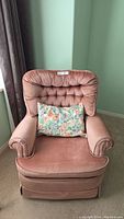 Front view of a pink velour arm chair with button tufted backrest and floral cushion on seat.