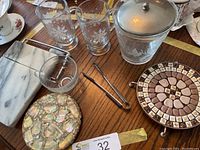 Glass ice bucket with lid, two etched glass mugs, round marble cheese board, clear glass bowl, metal tongs, and mosaic inlaid trivet on a wooden table
