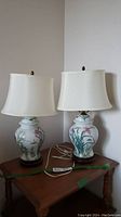 Two Asian style porcelain bedside lamps on wooden table, one with floral motif and the other with bird motif, both with off-white fabric lampshades and wooden bases.