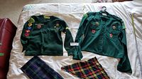 Full uniform laid out on bed including green scout shirt, sweater, and two plaid scarves