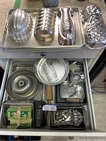 Top view of assorted metal baking molds on a tray plus drawer opened showing metal cookie cutters, pans, measuring cups, and other baking tools.