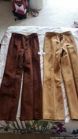Two pairs of custom made suede pants on a bed. One pair dark brown, the other tan. Both pants have button and zipper closures visible from front view.