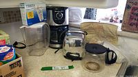 KitchenAid coffee maker with glass pot, Brita water filter jug with box of 3 replacement filters, Sunbeam toaster with cover and extra coffee pot, along with Braun accessories and coffee filters arranged on a kitchen counter.