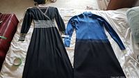 Two full-length pure wool sweater dresses laid flat on bed, black dress with striped waist and cuffs left, blue and black dress with zipper right.