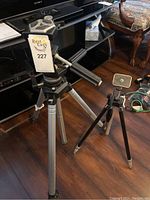 Two tripods placed near a TV stand on wooden flooring. One tripod has a label 'Rest Easy 227' attached to it and is significantly larger with black handles and aluminum legs.
