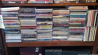 Wooden shelf displaying several rows of standard-size and slim jewel-case audio CDs stacked horizontally.