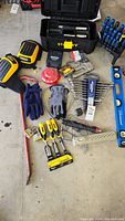 Overall assortment of hand tools arranged on garage floor including Stanley toolbox, gloves, wrenches, screwdrivers, chisels.