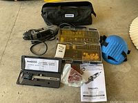 Photo showing Dremel rotary tool, accessory kit, carrying bag, electronic caliper in case, instruction manual, and blue knee pad on a concrete floor.
