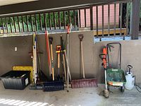 Photo showing nine garden tools including shovels, rakes, a snow shovel, and a weed sprayer along with a black plastic bin on the ground.