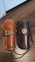 Two leather holsters side by side, showing front details including stitching, tooling, buckle, and sheriff star badge on dark holster.