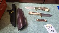 Four knives and two sheaths laid out on carpet: two fixed blades with stag handles, one pocket knife with stag scales, one smaller fixed blade, brown leather sheath and black nylon sheath