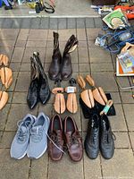 Lot of various men's footwear and wooden shoe stretchers on outdoor pavement; includes sneakers, boots, boat shoes, brogues, and several wooden shoe stretchers.