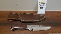 Knife with wooden handle and patterned blade alongside brown leather sheath with Knife King logo.