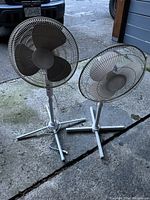 Two standing floor fans with adjustable poles and metal blades standing on concrete ground outdoors.
