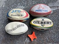 Four rugby balls including a white Commander ball, a Gilbert ball with yellow and green accents, a ball printed with 'Jump.ca' and SaskTel logos, and a ball labeled 'Practice' featuring an image of players. An orange plastic kicking tee with a wide base and V-shaped holder.