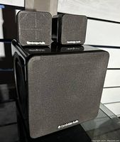 Front view of Cambridge Audio subwoofer with two satellite speakers displayed on top, showing fabric-covered fronts and branding.