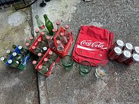 Photo showing multiple vintage glass soda bottles in plastic crates (Coca-Cola and Sprite), Coca-Cola branded red backpack, collectible soda cans, and an assortment of glassware.