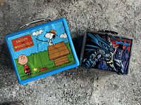 Photo shows two collectible lunch boxes side by side on a concrete background. The left is a Snoopy and Woodstock themed lunch box with cartoon art, and the right is a Batman lunch box with a three-dimensional raised Batman figure and stylized city background.