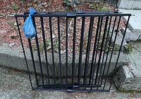 Two black metal adjustable toddler/pet gates leaning against concrete steps with installation hardware attached in plastic bag.