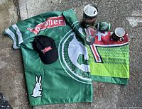Overview photo showing a large green Saskatchewan Roughriders flag, a black Pilsner cap, a Pilsner tin canister, two flags including a red and green one, a glass pint, a green miniature football, and a green ceramic mug.