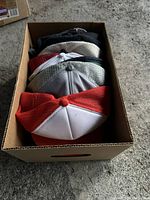 Top view of multiple trucker hats stacked in a cardboard box showing colors red, white, gray, and black with mesh backs.
